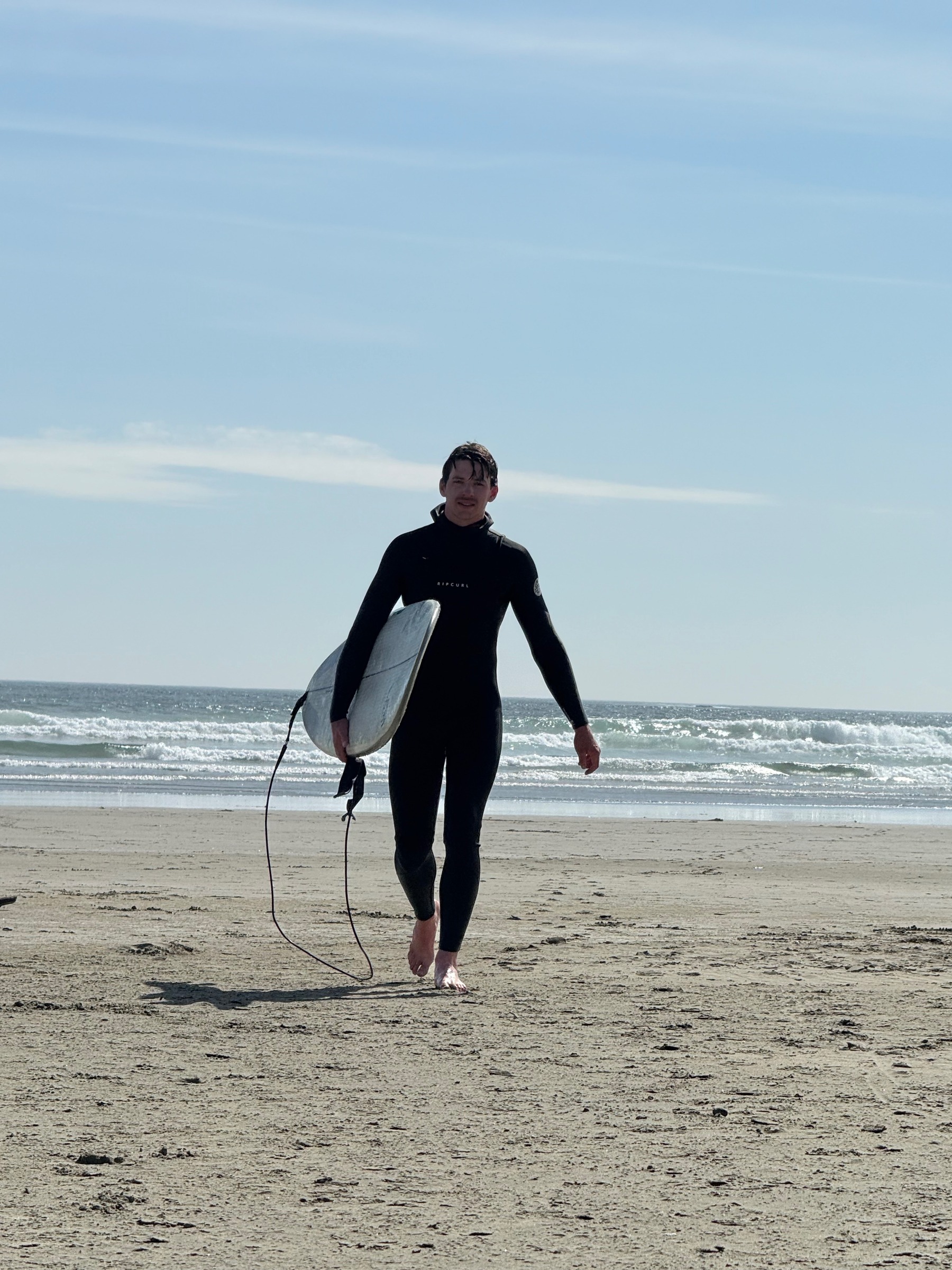 Everett Procter, professional athlete from Vancouver Island, walking on the beach in a wetsuit carrying a surfboard